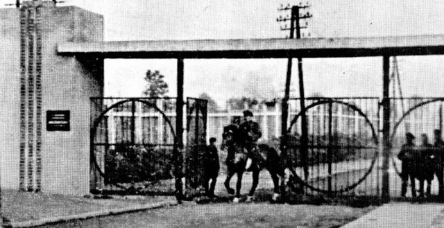 Black and white photo of a camp entrance gate with a Nazi eagle emblem, men in uniform, and circular metalwork.