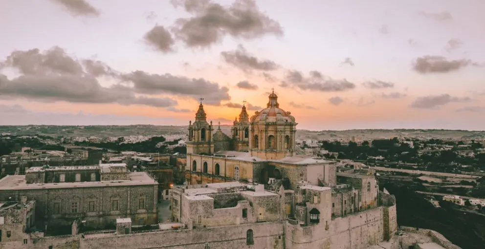 Aerial view of Mdina's St. Paul's Cathedral with domes and bell towers, surrounded by fortified walls and historic buildin...