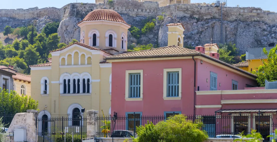 Athens cityscape: A yellow church and pink building in the foreground, with the Acropolis and Parthenon in the background.