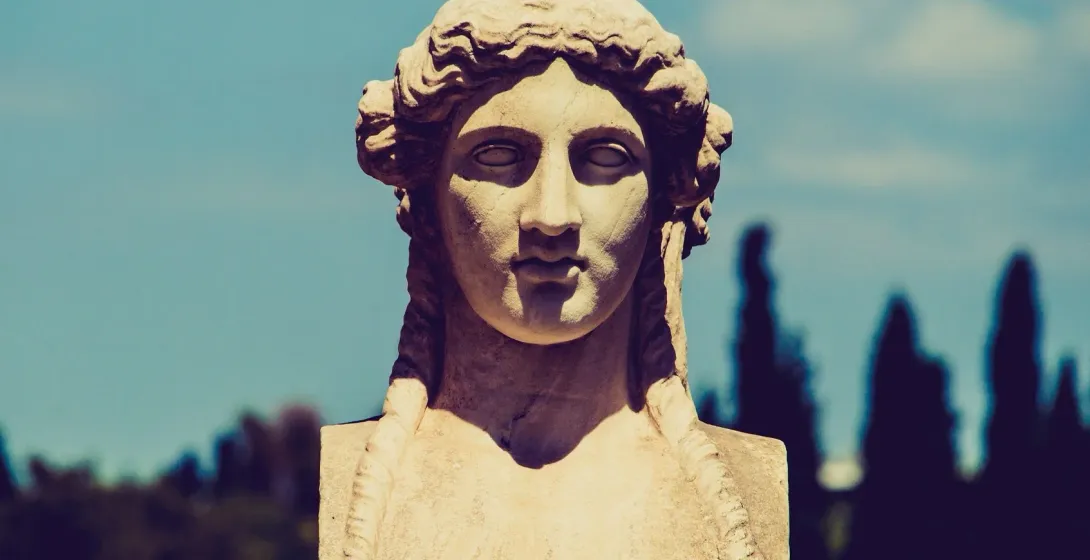 Close-up of an ancient Greek marble bust of a woman with braided hair, against a clear blue sky.