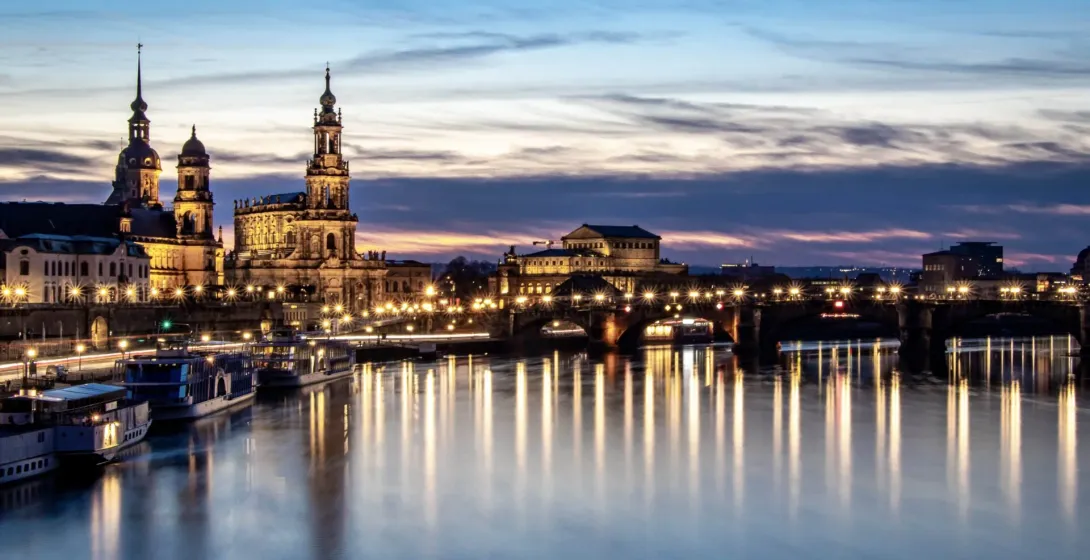 Illuminated Dresden skyline at dusk with Frauenkirche, Hofkirche, Augustusbrücke bridge, and boats on the calm Elbe River.