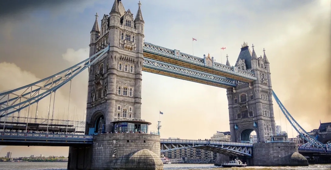 Tower Bridge spanning a wide river under a partly cloudy sky, with a boat on the water and buildings in the distance.