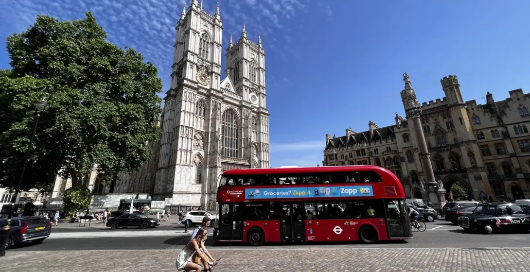 Westminster Abbey's historic facade and tower under a blue sky, with a red double-decker bus, cars, and people on the street.