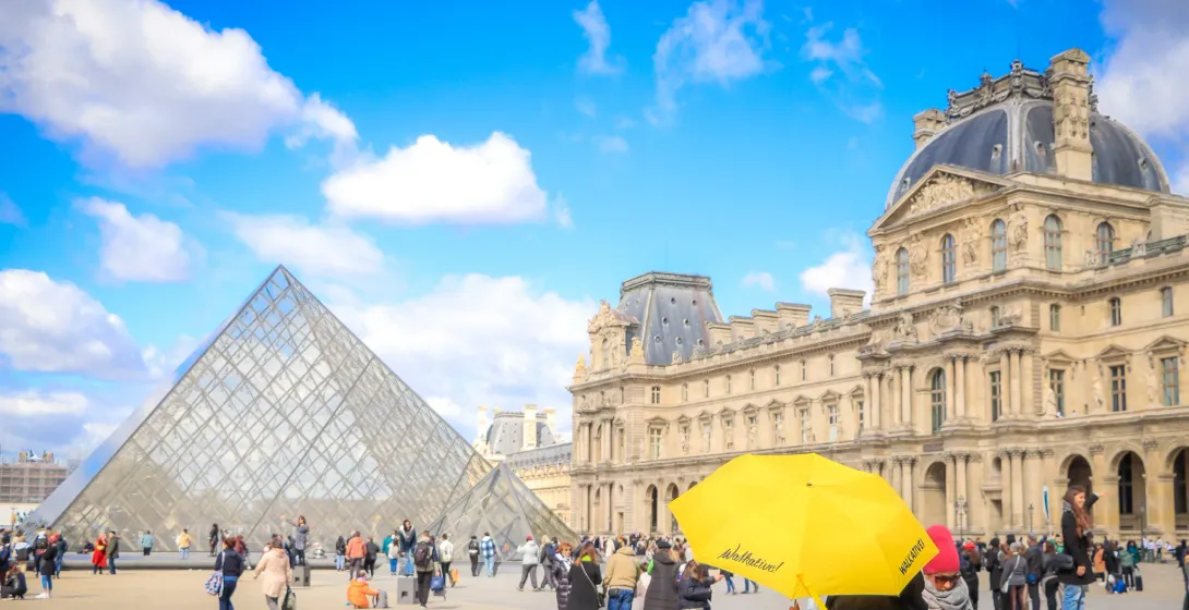 The glass pyramid entrance of the Louvre Museum on a sunny day with clouds, surrounded by classical palace buildings and v...