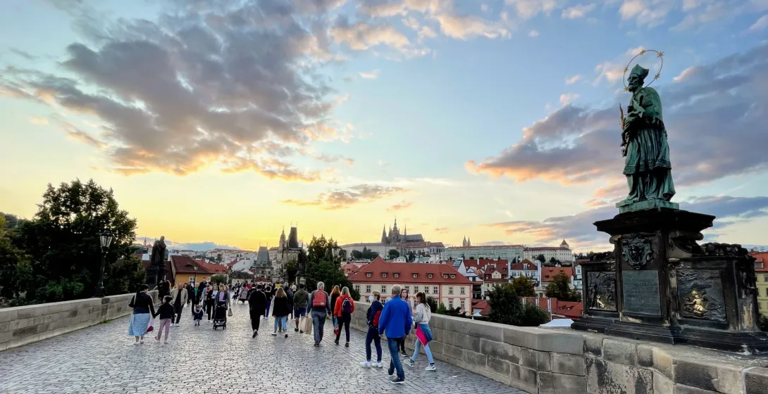 Tourists walking across Charles Bridge in Prague at sunset, with Prague Castle in the background.