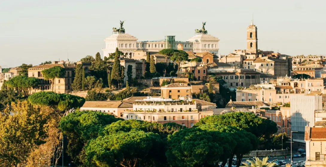 Panoramic view of Rome with the Victor Emmanuel II Monument atop a hill, a busy street with cars and buses, and green trees.