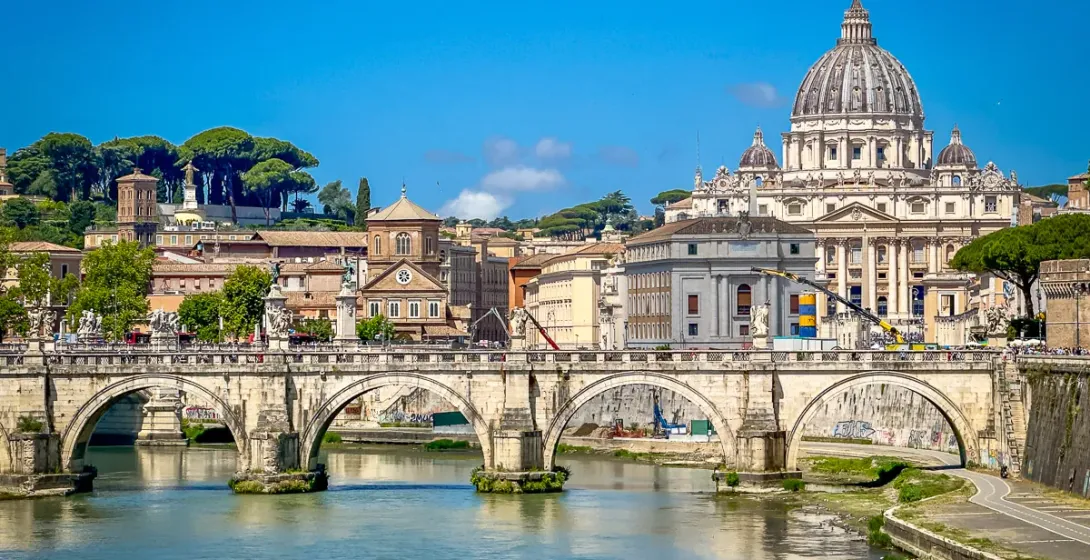 Sant'Angelo Bridge over the Tiber River, with St. Peter's Basilica dome and Roman buildings under a clear blue sky.