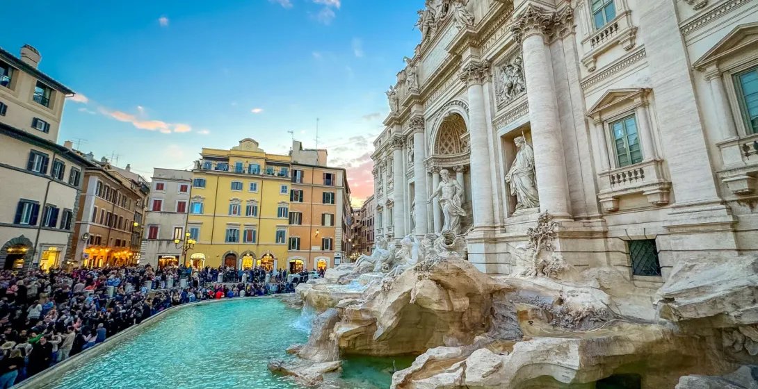 Ornate white Trevi Fountain in Rome with turquoise water, surrounded by a large crowd of people under a blue and pink sky.
