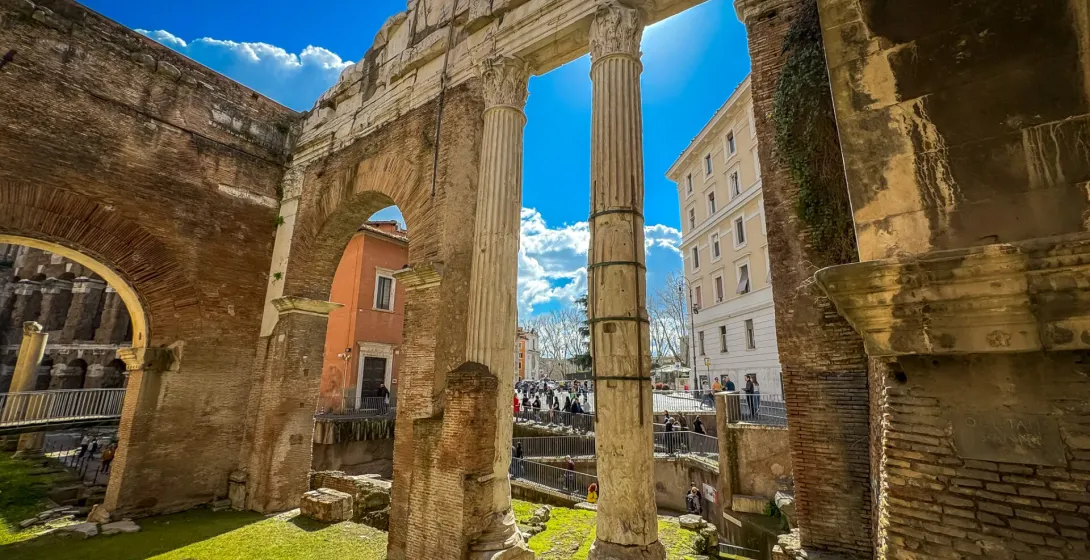 Ancient Roman brick ruins with arches and two tall stone columns. Green grass fills the excavated area, modern buildings v...