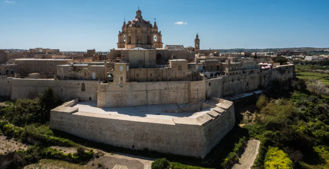 Aerial view of a historic fortified city with golden stone walls, buildings, and a large domed cathedral under a blue sky.