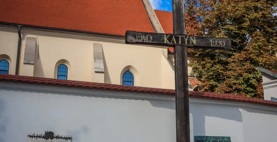 Large wooden Katyn Cross memorial with 'KATYN 1940 1990' inscription, plaques on a white wall, church roof, and spire.