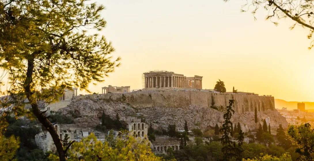The Acropolis of Athens with the Parthenon and scaffolding, bathed in warm sunset light, seen through green trees.