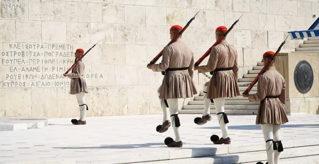 Four Evzones soldiers in traditional uniform marching in formation in front of a carved marble wall.