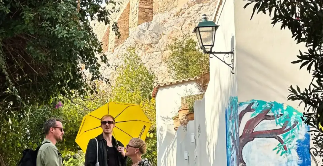 Three people on stone stairs in Athens, one with a yellow umbrella. A building with a tree mural and a large stone wall ar...