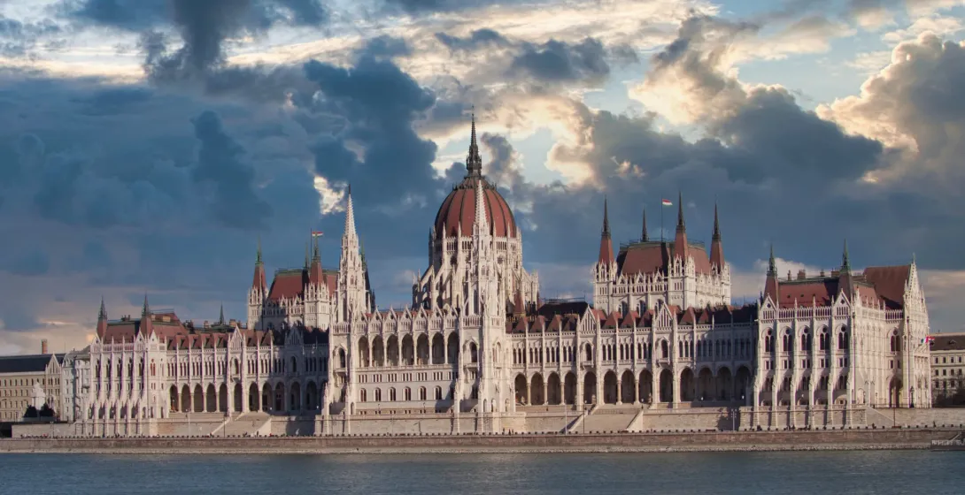 The Hungarian Parliament Building viewed from across the Danube River under a dramatic, cloudy sky.