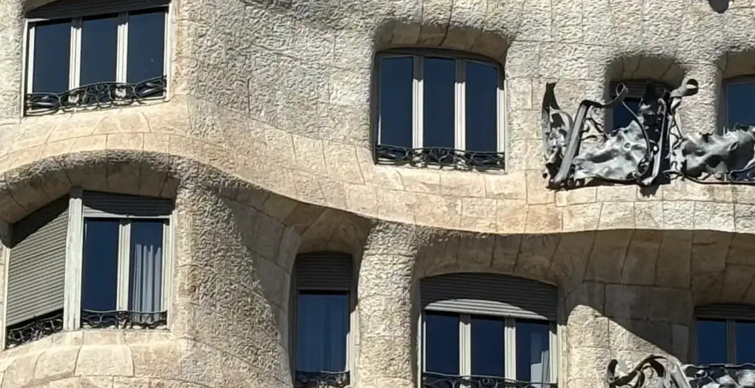 Close-up of Casa Milà's wavy stone facade with dark windows, grey roller blinds, and wrought-iron balconies under a blue sky.