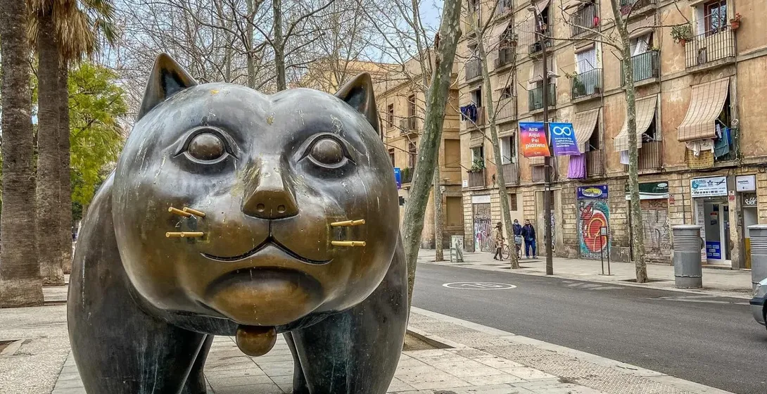 Large bronze sculpture of a plump cat with small ears, on a city sidewalk next to a street with buildings and trees.