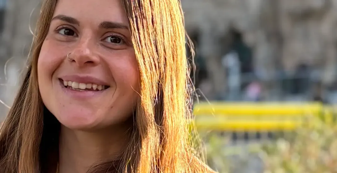 Smiling woman with long brown hair, brown coat, and yellow 'Catalonia Tourism Guide' lanyard. Blurry building and palms in...