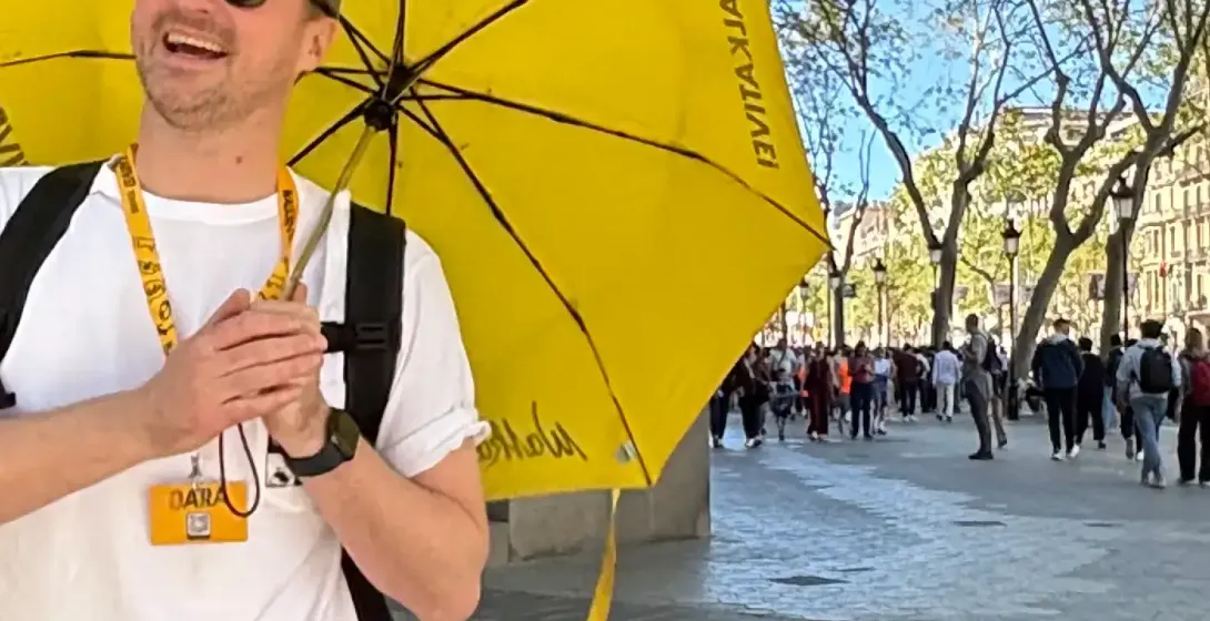 A smiling male tour guide holding a yellow Walkative umbrella on a sunny street in Barcelona. Ornate modernist building in...
