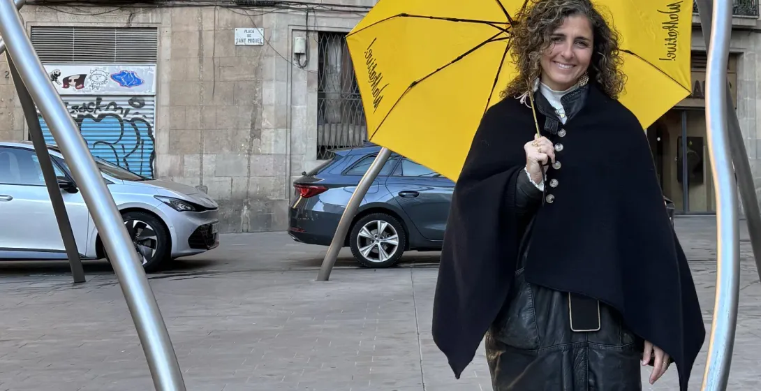 Smiling woman with curly hair holds a yellow Walkative umbrella in Plaça de Sant Miquel, Barcelona. Buildings and cars beh...