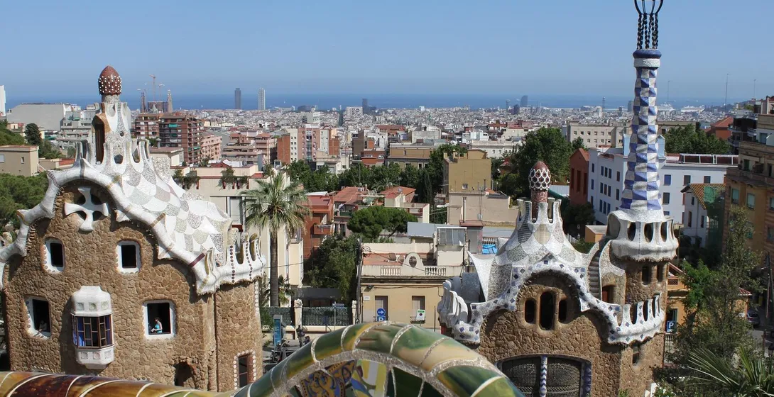 View from Park Güell showing two Gaudi-designed mosaic-roofed buildings, a colorful tiled wall, and Barcelona cityscape wi...