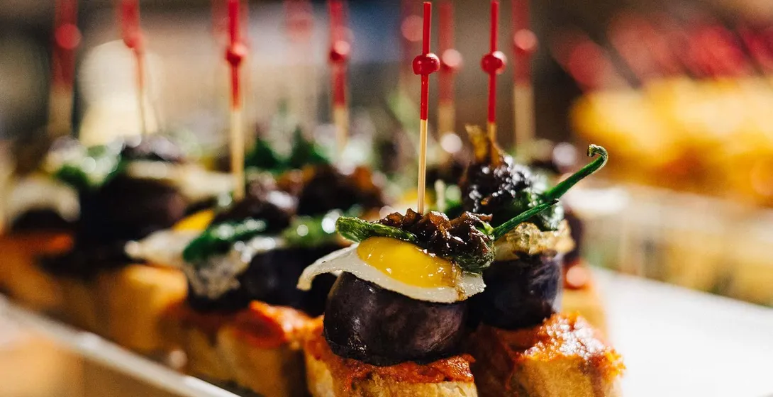 Close-up of a plate of pintxos, small open-faced sandwiches with various toppings, held by red toothpicks. A dark sausage ...