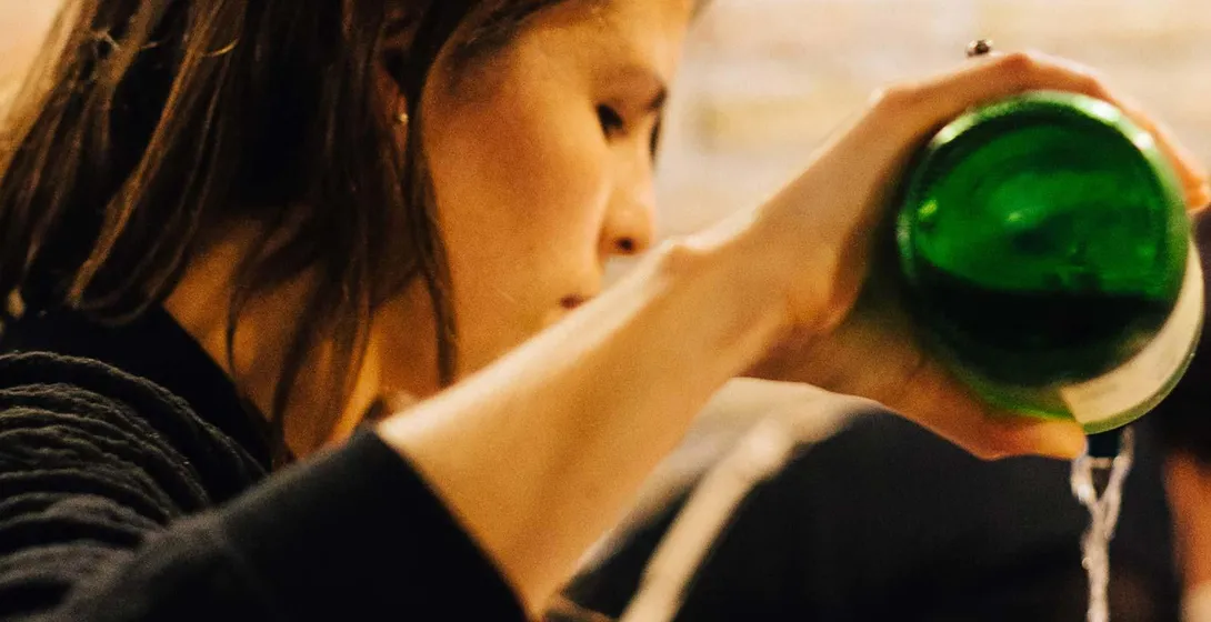 Person pouring sidra from a green bottle into a glass, generating foam. Blurred stone wall in background.