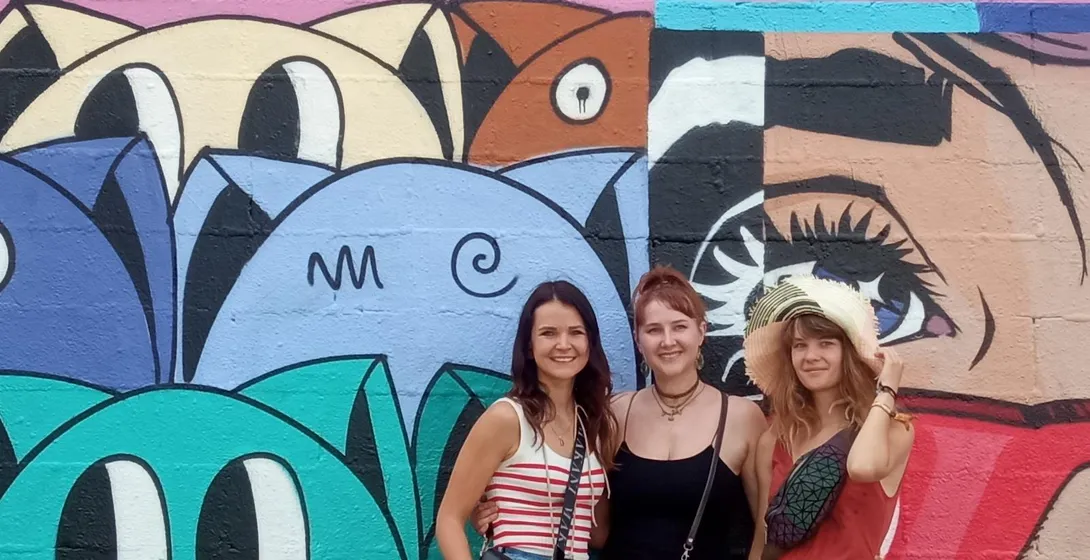 Three women smiling in front of a colorful wall covered in abstract graffiti and a large mural with stylized faces.