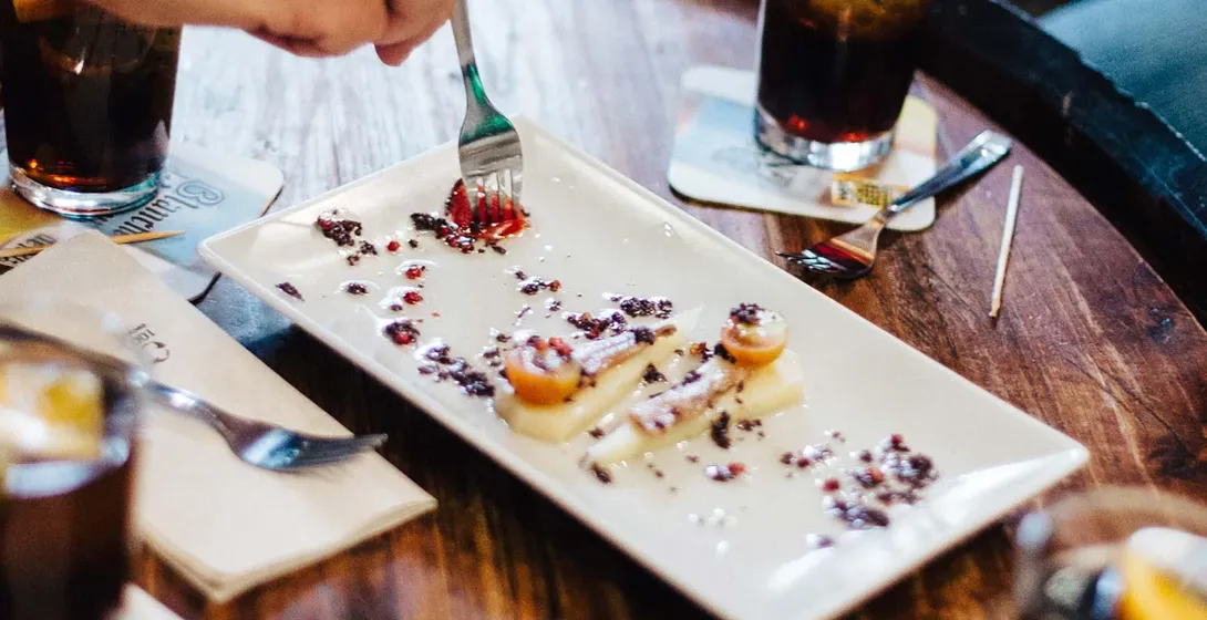 People's hands around a wooden table with tapas plates and drinks. One person uses a fork to eat a dish.