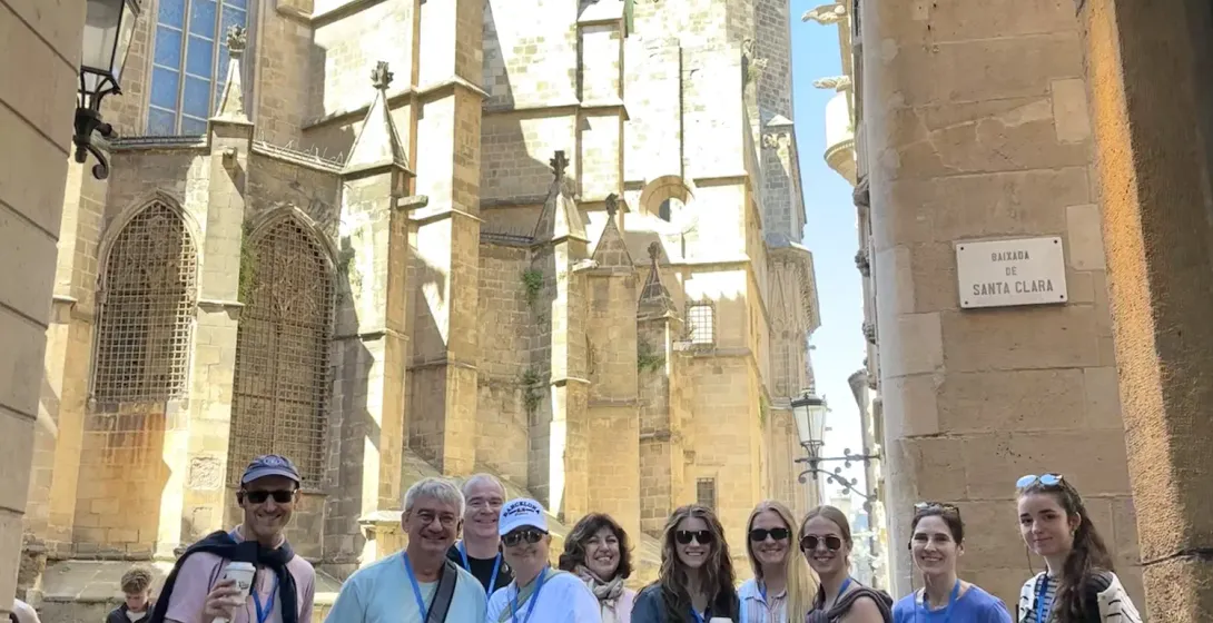 A diverse group of people stands on a narrow stone street with Barcelona Cathedral in the background under a blue sky.
