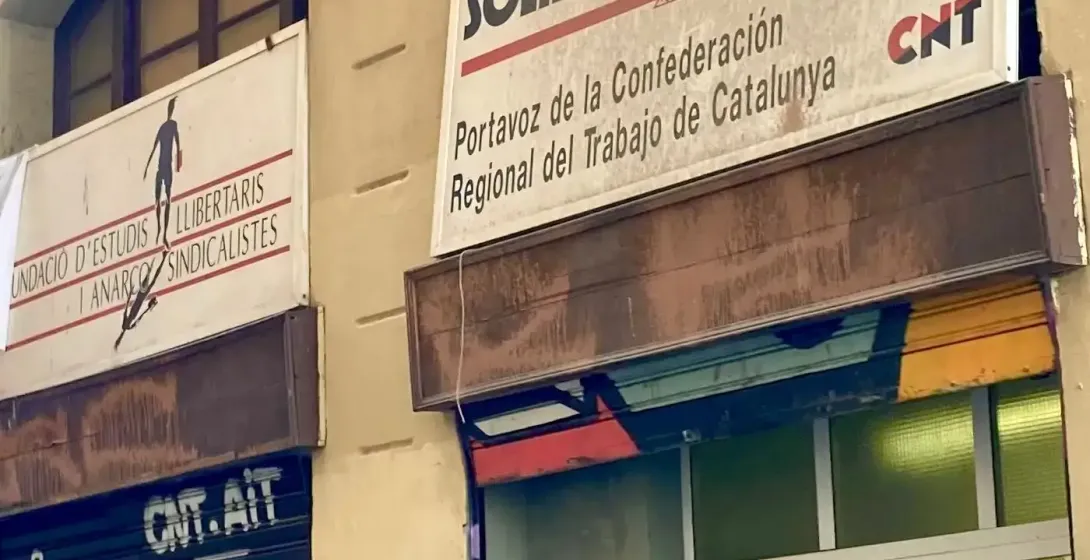 A group of people on a street tour looking up at a building with "Solidaridad Obrera" and "CNT" signs, and graffiti on a s...