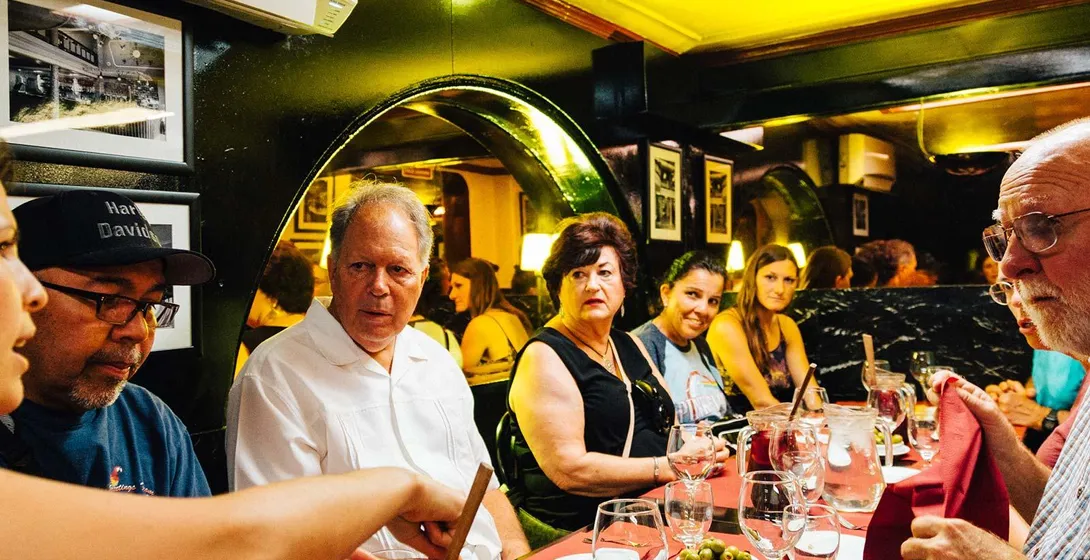 A group of people sitting at a restaurant table with red tablecloths, sharing tapas dishes, olives, and a pitcher of red s...