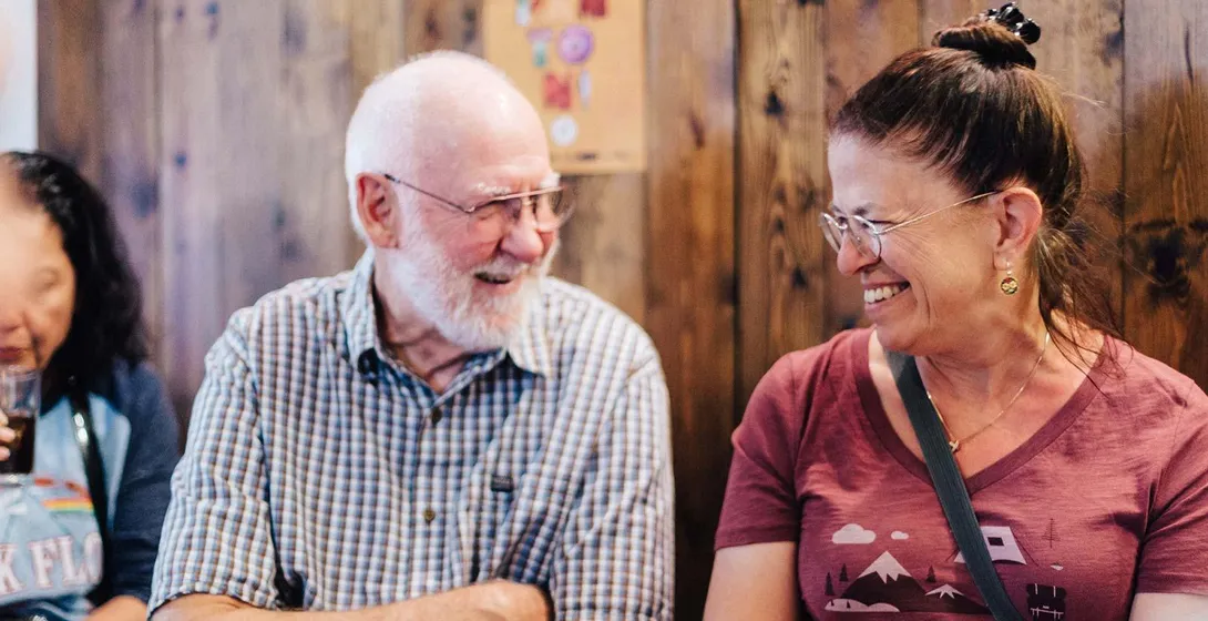 Two smiling travelers, an older man and a woman, talking in a casual setting with a wooden wall and a tapas poster.