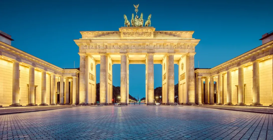 The Brandenburg Gate in Berlin, illuminated at dusk, featuring the Quadriga sculpture on top and a wide, empty cobblestone...