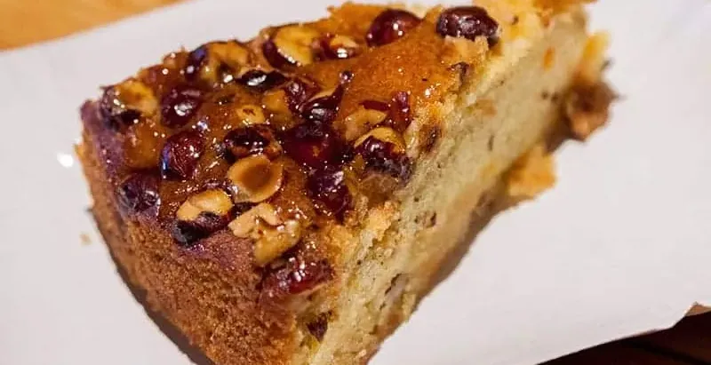 Slice of cake with a glossy topping of roasted hazelnuts and dried berries on a white plate.