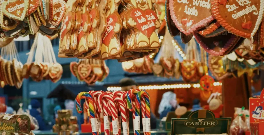 A stall at a Christmas market selling gingerbread hearts, candy canes, and roasted nuts under warm lights.