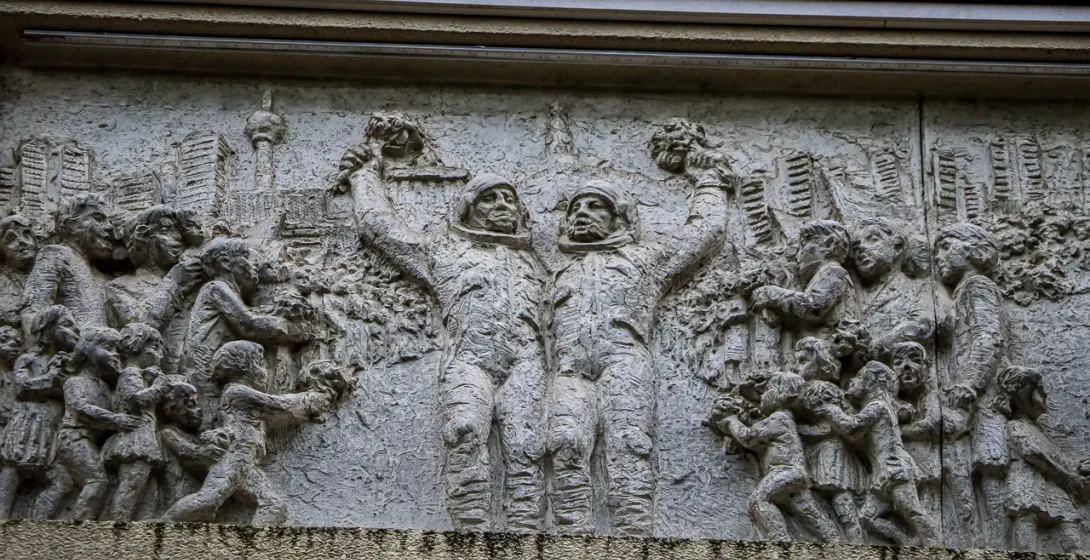 Detailed stone relief sculpture depicting two cosmonauts holding bouquets, surrounded by adults and children, with the Ber...