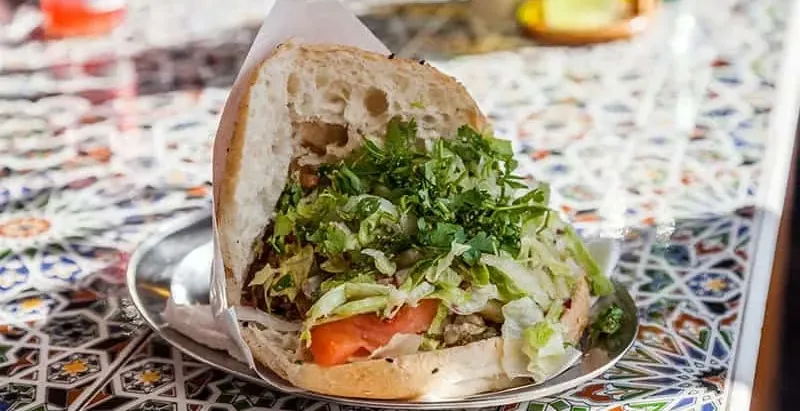Half a doner kebab in pita bread with meat, lettuce, parsley, and tomato, served on a metal plate on a tiled table.
