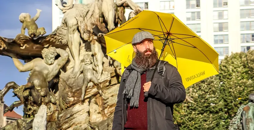 Bearded man in a hat holding a yellow Walkative umbrella, standing in front of the Neptune Fountain in Berlin.