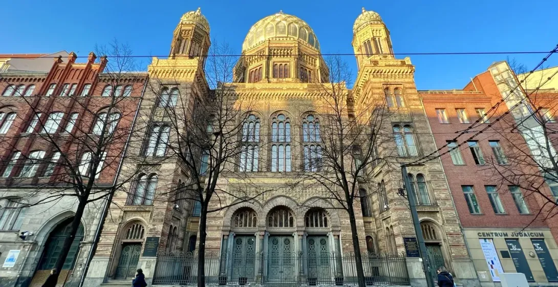 The ornate facade of the New Synagogue in Berlin on a sunny day, viewed from across the street through bare trees.
