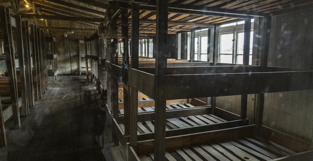 Interior of a wooden barrack with multi-tiered bunk beds, long rows extending into the dimly lit space. Windows are visibl...
