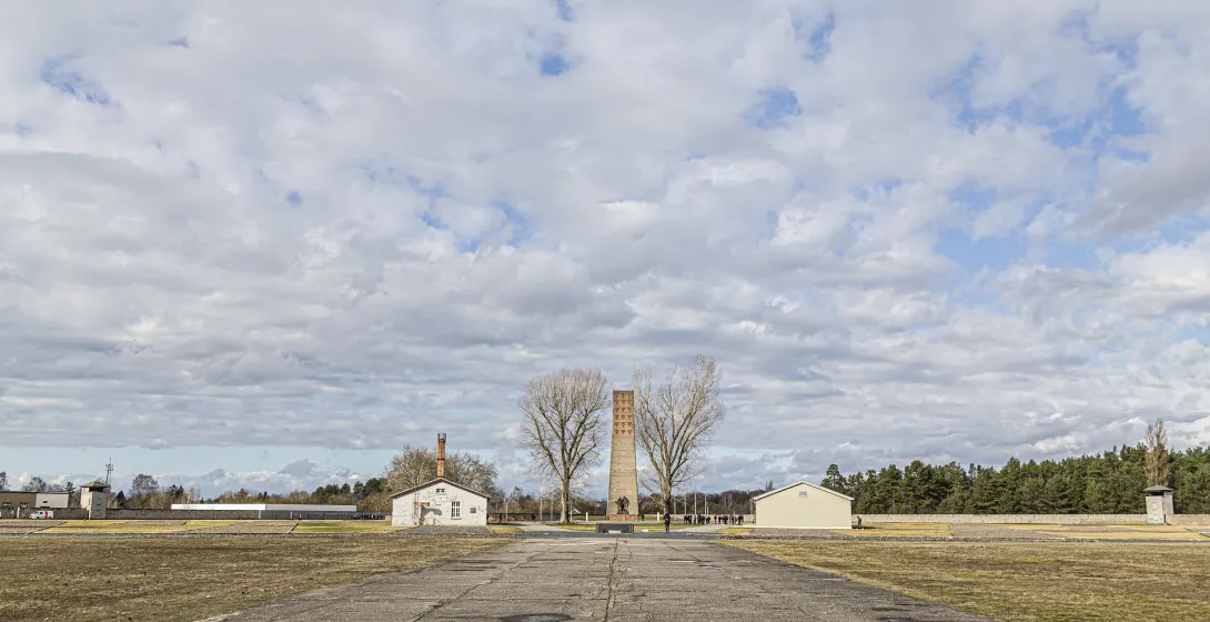 Concrete path leading to a tall, triangular brick monument flanked by two bare trees, with low buildings and fields under ...