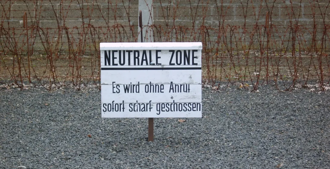 White sign with "NEUTRALE ZONE" and warning text in German, in front of barbed wire fence and concrete wall on gravel ground.