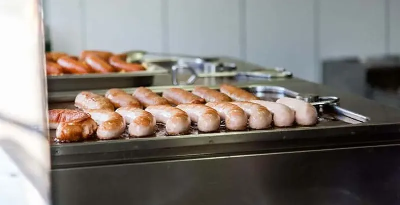 Sausages cooking on a metal griddle in a kitchen setting with white tiled walls. Metal tongs are visible.