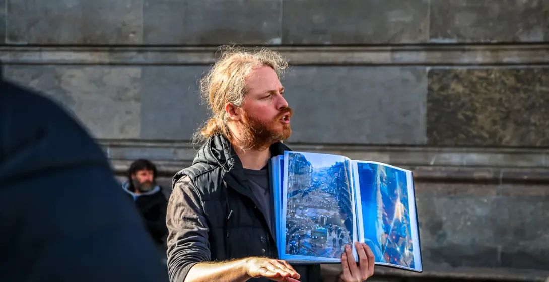 A tour guide in Berlin holds an open book showing a historical photo of a destroyed street while speaking to a group.