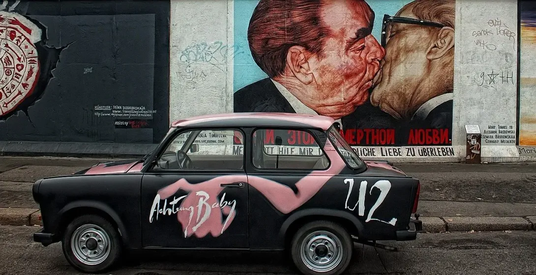 A black Trabant car parked in front of the 'Fraternal Kiss' mural on the East Side Gallery in Berlin.