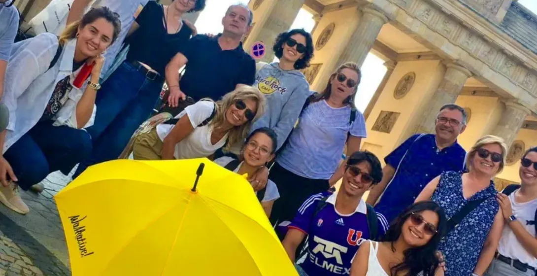 A diverse group of people posing with a yellow Walkative umbrella in front of the Brandenburg Gate in Berlin.