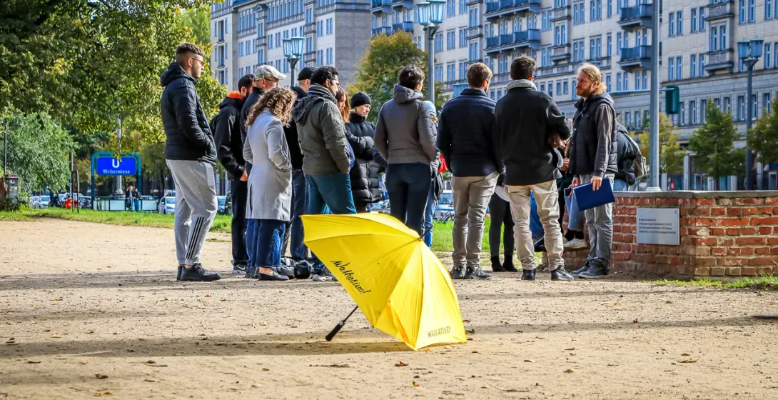 A group of people listening to a guide near apartment buildings in Berlin. A yellow Walkative umbrella rests on the ground.