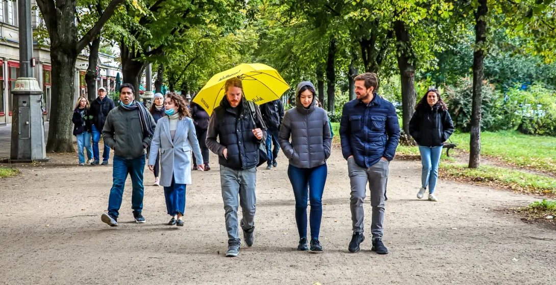 Group of people walking on a wide dirt path lined with mature trees. Buildings are visible on the left, one person holds a...