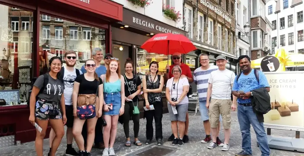 A diverse group of tourists stands smiling on a cobblestone street in front of Belgian Lace and Chocolate shops.